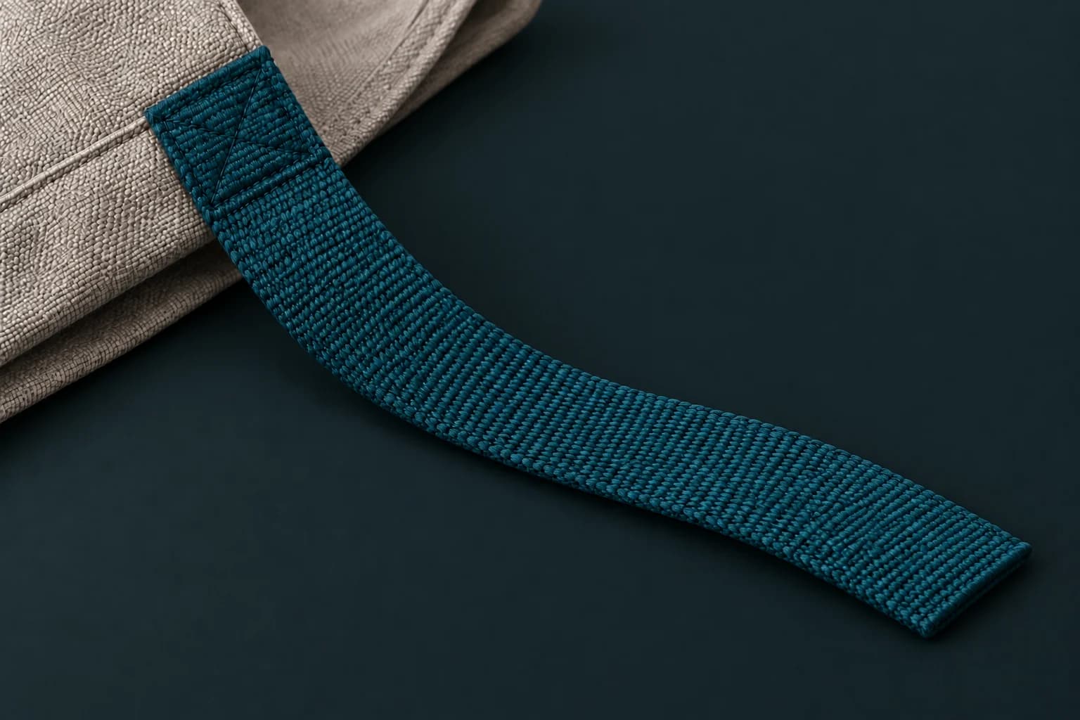 A teal flat woven strap stitched onto grey bag fabric with its free end visible